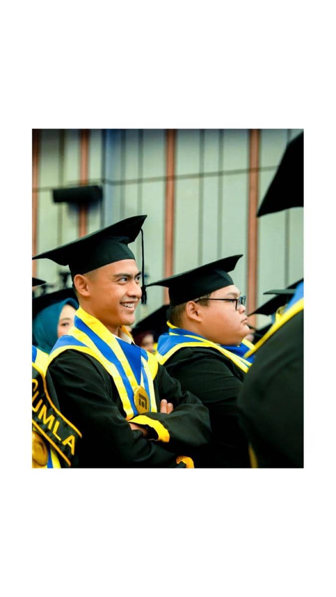 Pratama Arhan mengenakan toga dan selempang biru kuning tersenyum saat menghadiri upacara kelulusan di aula kampus.