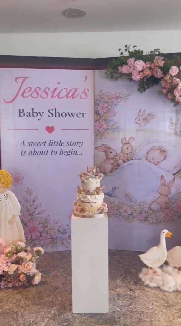 Dekorasi baby shower bertema pastel dengan kue dua tingkat, bunga mawar merah muda, dan latar bergambar kelinci serta angsa.