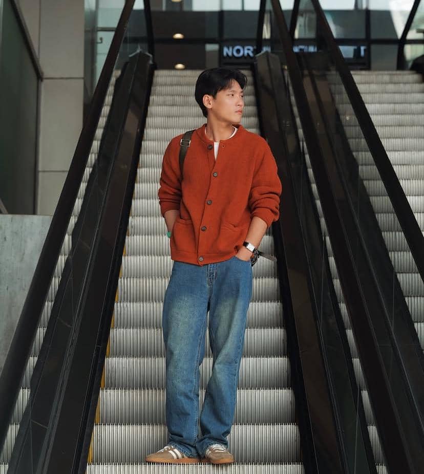 Seorang pria mengenakan cardigan oranye dan celana jeans berdiri di eskalator dalam gedung modern sambil menatap ke samping.