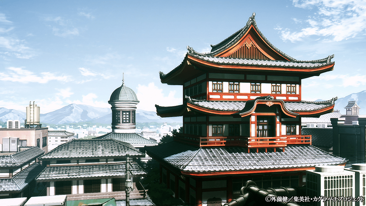 Bangunan bergaya tradisional Jepang dengan atap bertingkat merah dan abu-abu di tengah kota, berlatar pegunungan dan langit cerah.