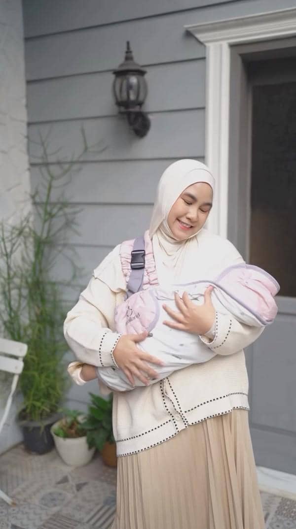 Seorang wanita berhijab tersenyum sambil menggendong bayi yang dibungkus kain berwarna lembut di depan rumah berwarna abu-abu.