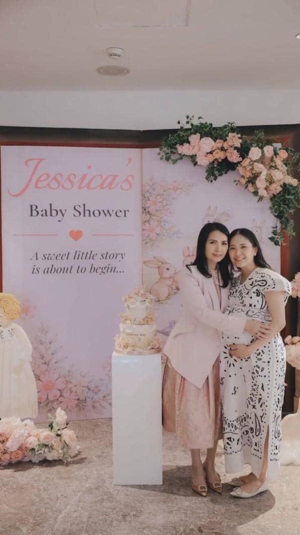 Dua wanita berpose di acara baby shower bertema pastel dengan dekorasi bunga dan kue bertingkat di depan latar bertulisan Jessica's Baby Shower.