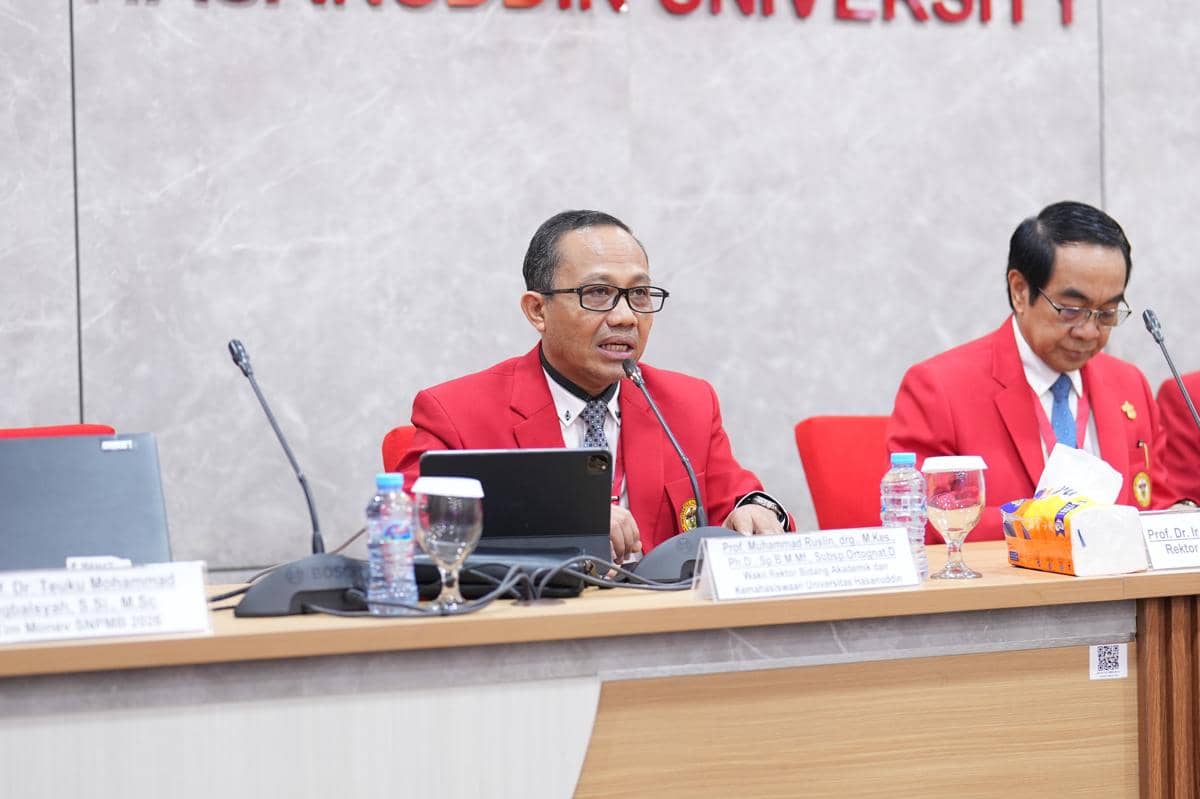Seorang pria mengenakan jas merah berbicara di depan mikrofon dalam sebuah rapat di Universitas Hasanuddin bersama rekan sejawatnya.