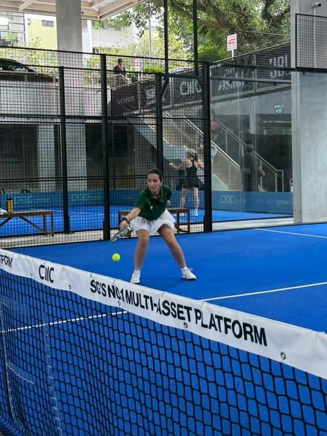 Seorang perempuan mengenakan kaus hijau dan celana putih sedang memukul bola dalam pertandingan padel di lapangan biru di Singapura.