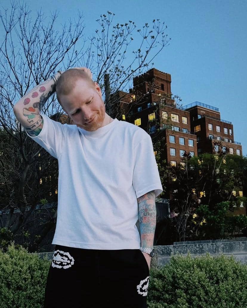 Seorang pria berambut pendek mengenakan kaus putih dan celana hitam berdiri di luar ruangan dengan latar gedung bata merah dan langit biru.