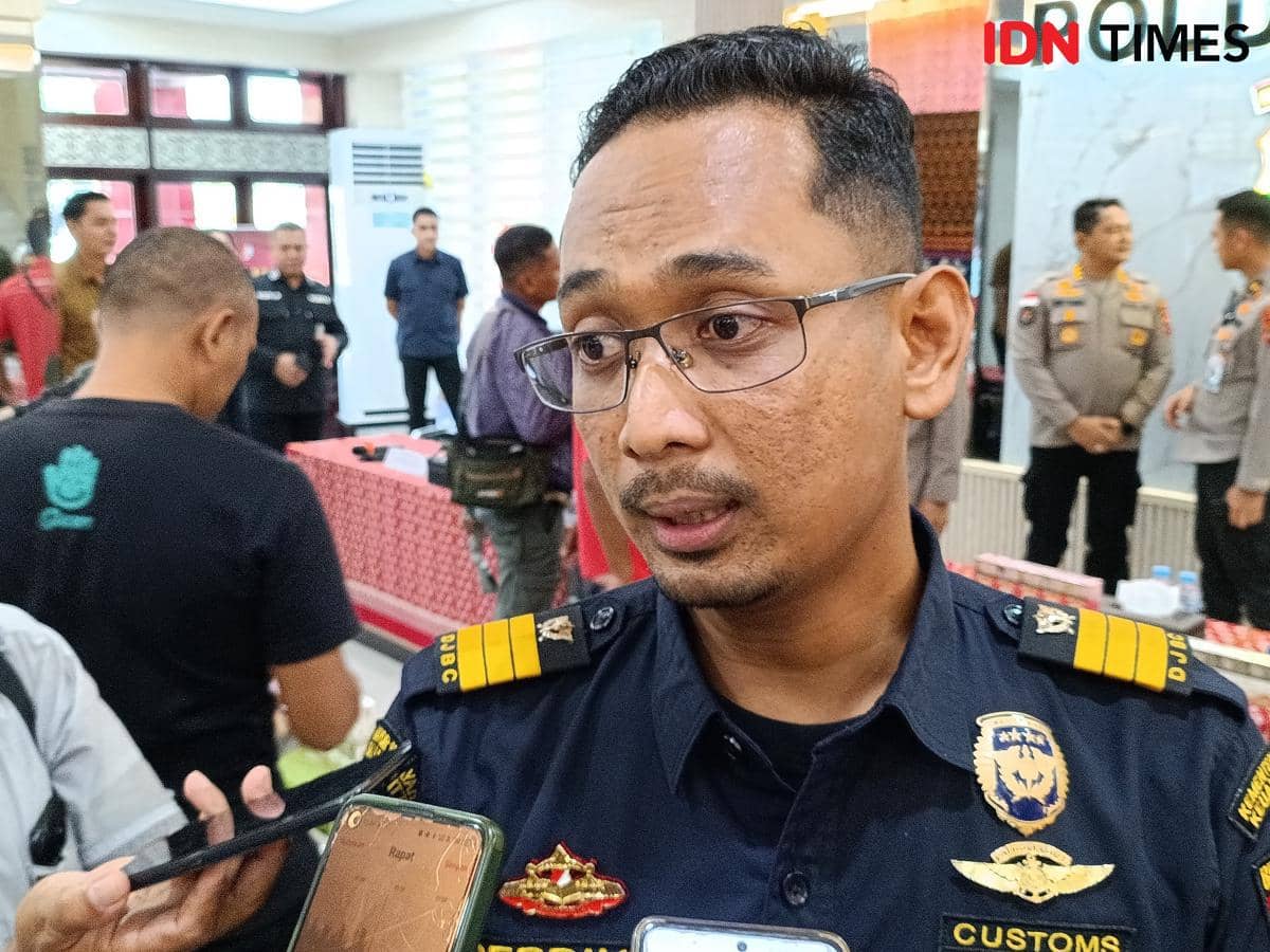 Seorang petugas Bea Cukai berseragam hitam dengan atribut resmi sedang diwawancarai oleh beberapa jurnalis di sebuah ruangan.