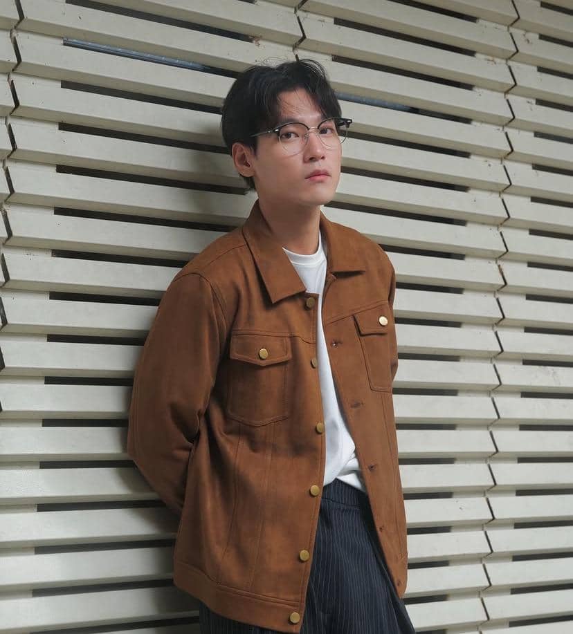 Seorang pria berjaket cokelat dan berkacamata berdiri bersandar di dinding berpola horizontal berwarna krem.