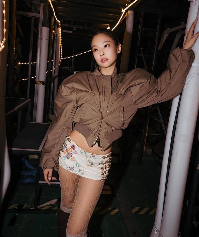 Seorang perempuan mengenakan jaket cokelat dan rok mini bermotif bunga berpose di area belakang panggung dengan pencahayaan lampu hias.