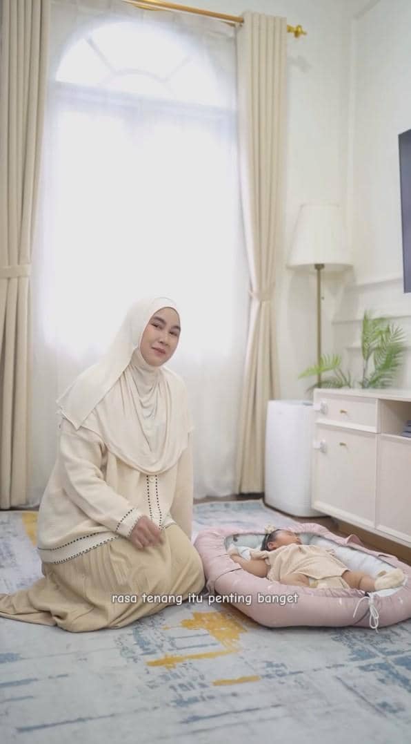 Seorang wanita berhijab duduk di lantai dekat bayi yang berbaring di tempat tidur bayi berwarna pink di ruang tamu bernuansa krem.