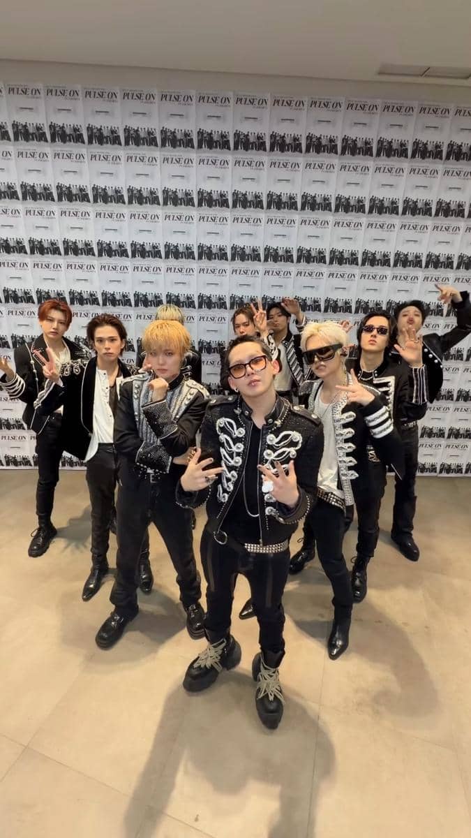 Grup beranggotakan sembilan orang berpose bersama mengenakan pakaian hitam dengan detail perak di depan dinding bertuliskan 'PULSE ON'.