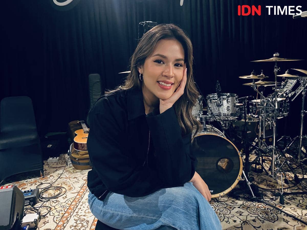 Seorang perempuan tersenyum sambil menopang dagu di studio musik dengan latar drum dan gitar di Jakarta.