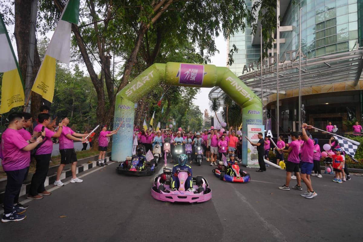 Peserta Fun Walk 5K Aryaduta Medan mengenakan kaus ungu muda bersorak di garis start dengan mobil go-kart berwarna cerah di depan hotel.