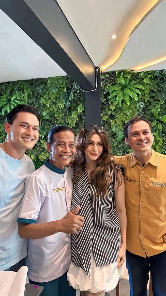 Empat orang berdiri berdekatan sambil tersenyum di depan dinding hijau berdaun, tampak akrab dan ceria dalam suasana hangat.