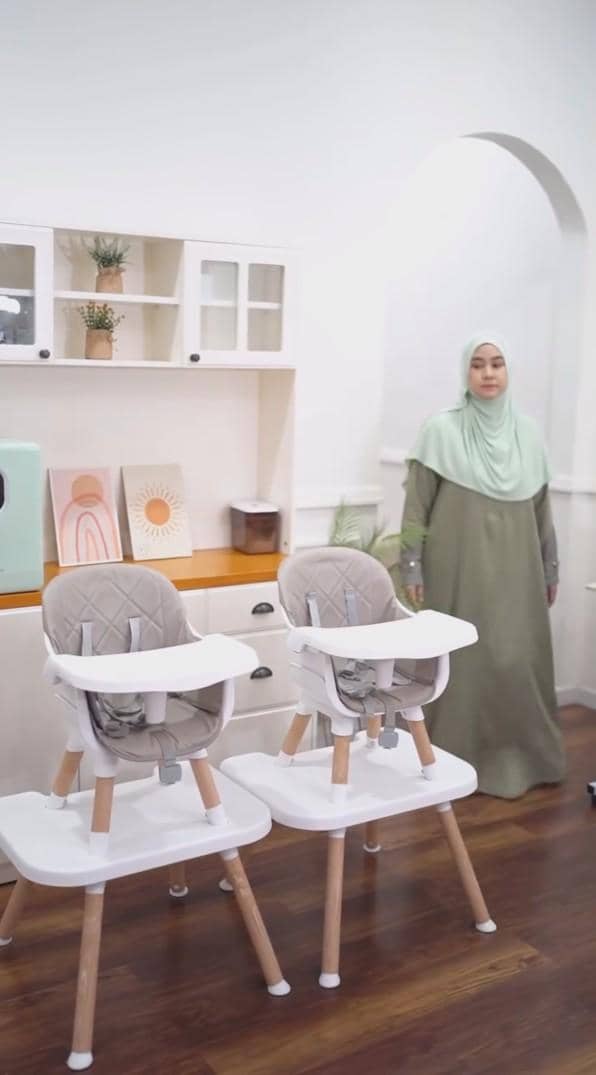 Seorang wanita berhijab berdiri di ruang makan minimalis dengan dua kursi bayi, rak putih, dan dekorasi bergaya Skandinavia.