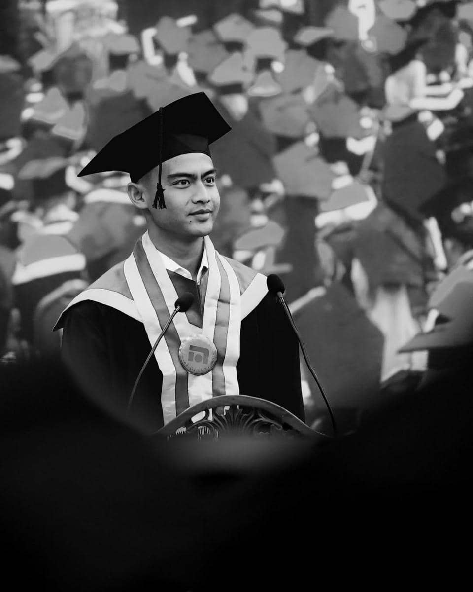 Pratama Arhan mengenakan toga dan medali berdiri di depan mikrofon saat upacara kelulusan dengan latar belakang para wisudawan lain.