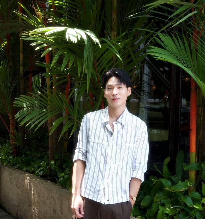 Seorang pria mengenakan kemeja garis-garis berdiri di luar ruangan dengan latar belakang tanaman hijau tropis yang rimbun.