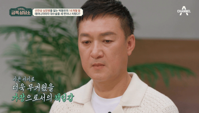 Seorang pria mengenakan sweter rajut putih dengan ekspresi serius di dalam ruangan dengan latar tanaman hijau.