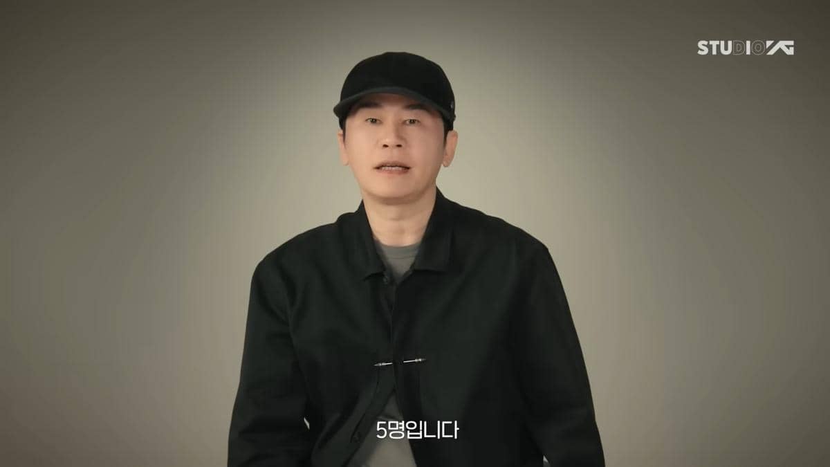 Seorang pria mengenakan jaket hitam dan topi hitam sedang berbicara di depan latar polos dengan logo Studio YG di pojok kanan atas.