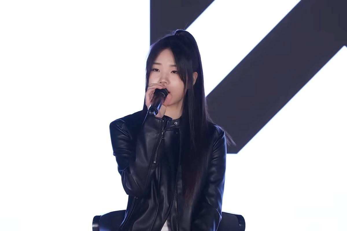 Seorang peserta pelatihan NEXT MONSTER mengenakan jaket kulit hitam sedang bernyanyi sambil memegang mikrofon di atas panggung.