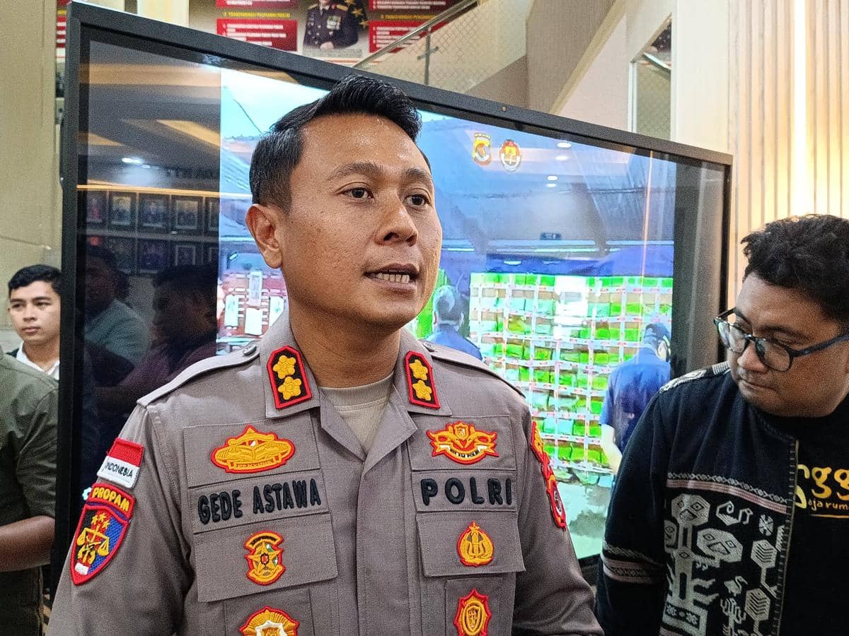 Seorang perwira polisi berpakaian dinas berbicara di depan layar besar dengan beberapa orang di sekitarnya di dalam ruangan.