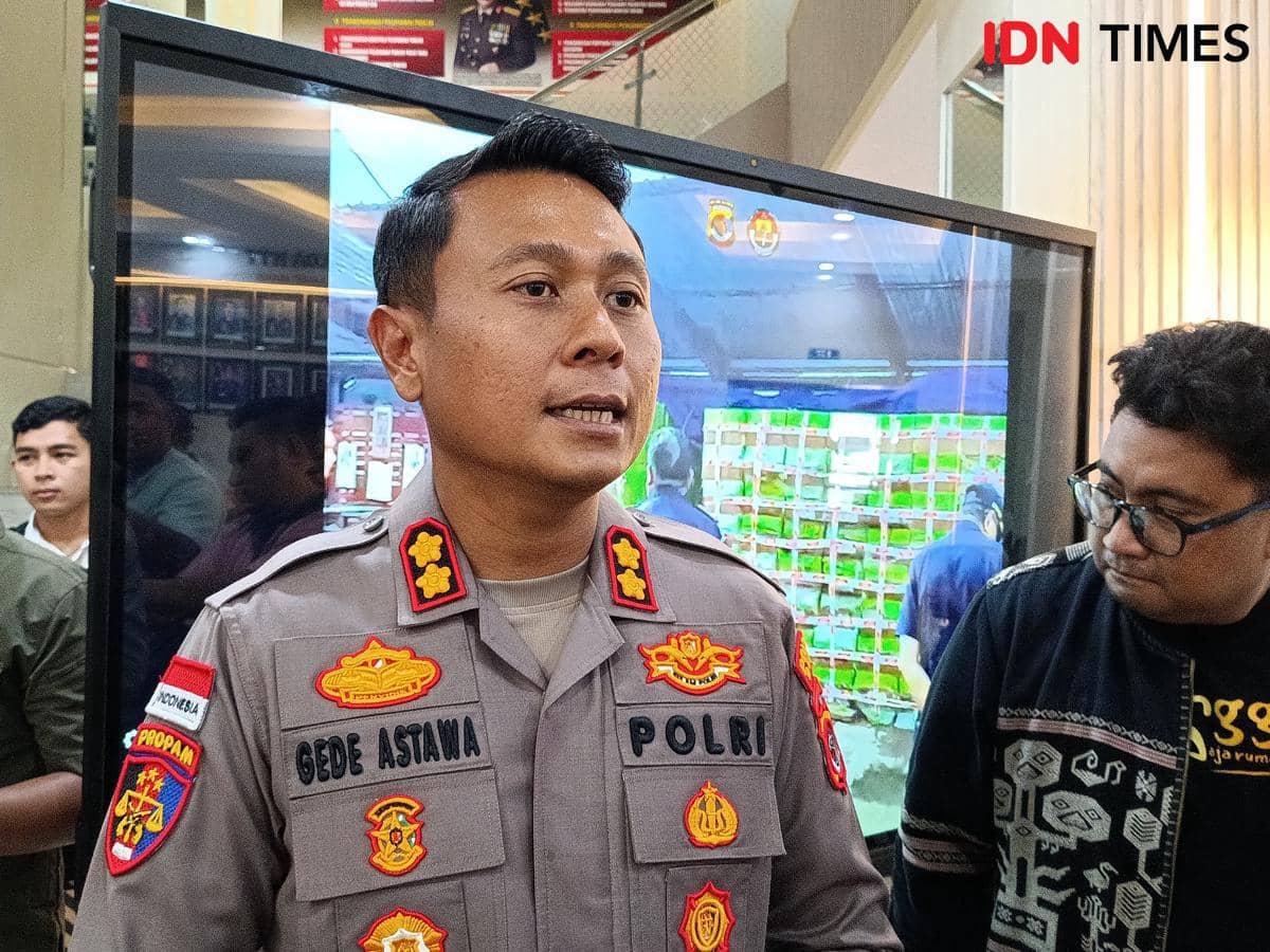 Seorang perwira polisi berpakaian dinas berbicara di depan layar besar dengan beberapa orang di sekitarnya di dalam ruangan.