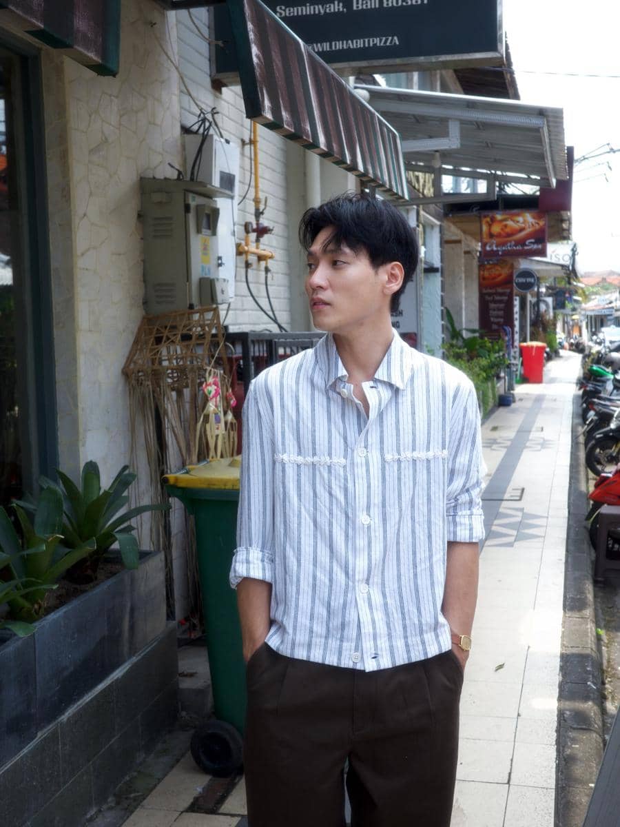 Na Daehoon mengenakan kemeja garis-garis biru putih dan celana cokelat berdiri di trotoar kawasan Seminyak, Bali.