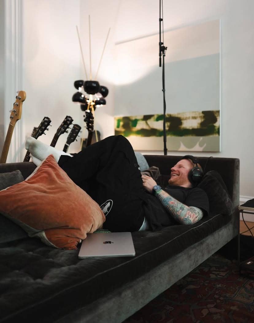 Seorang pria berbaring santai di sofa dengan headphone di telinga, dikelilingi beberapa gitar dan laptop di ruang bernuansa hangat.