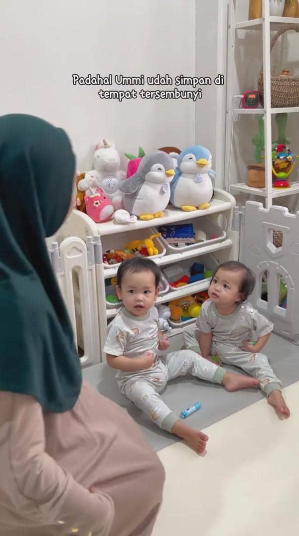 Dua balita kembar duduk di lantai ruang bermain dengan rak mainan berisi boneka dan mainan warna-warni di belakang mereka.