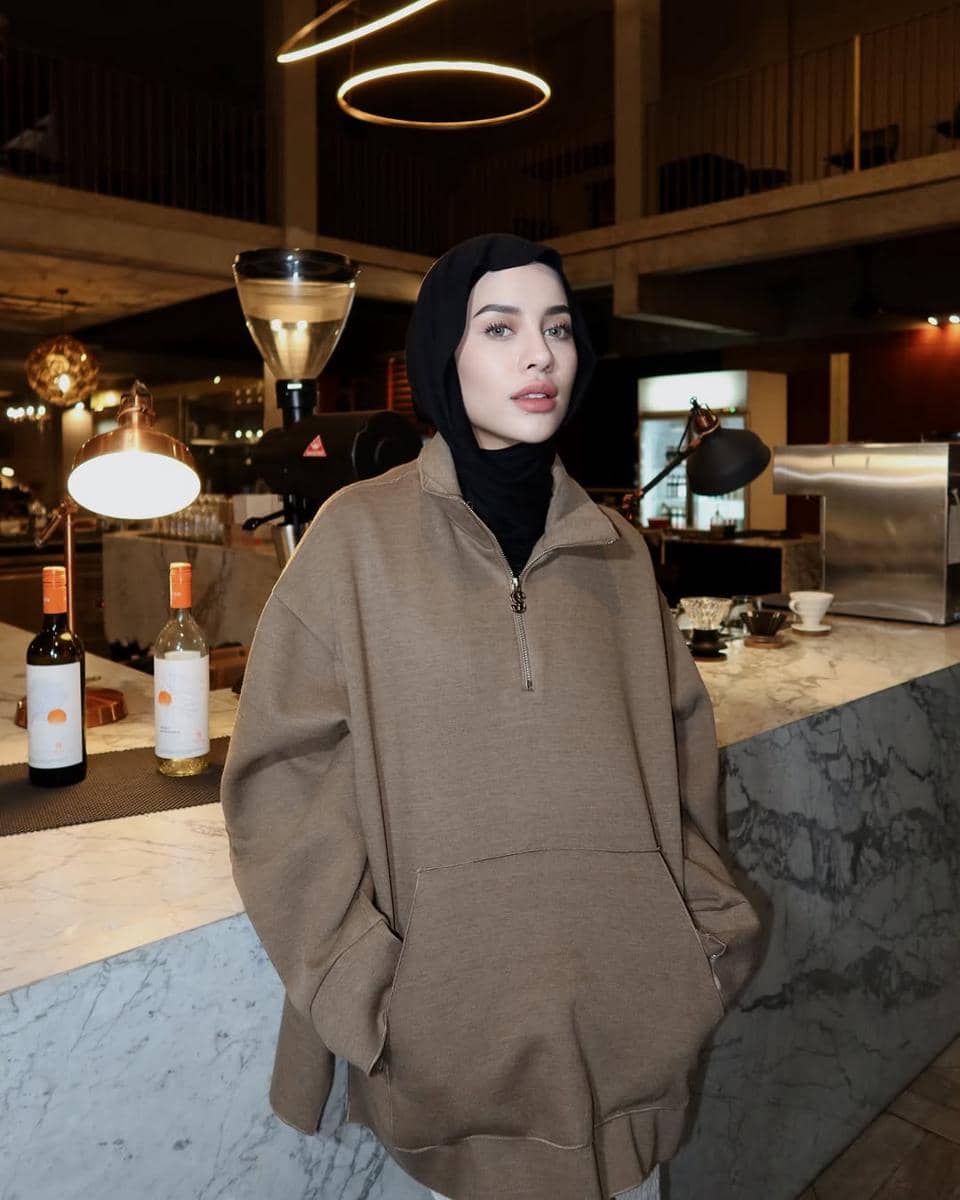 Seorang wanita berhijab hitam mengenakan sweater cokelat berdiri di depan meja marmer di kafe dengan pencahayaan hangat.