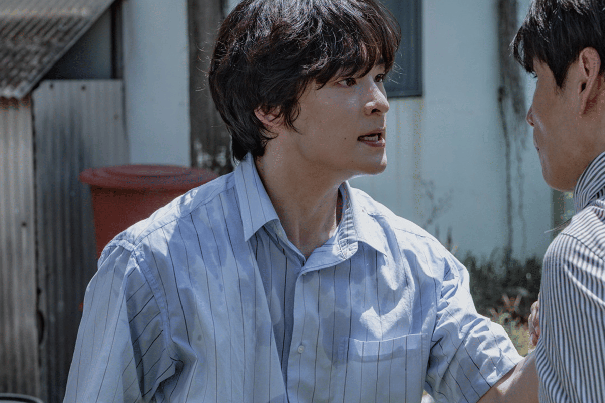 Seorang pria berwajah tegang mengenakan kemeja biru bergaris sedang memegang bahu pria lain di luar ruangan pada adegan drama The Scarecrow.