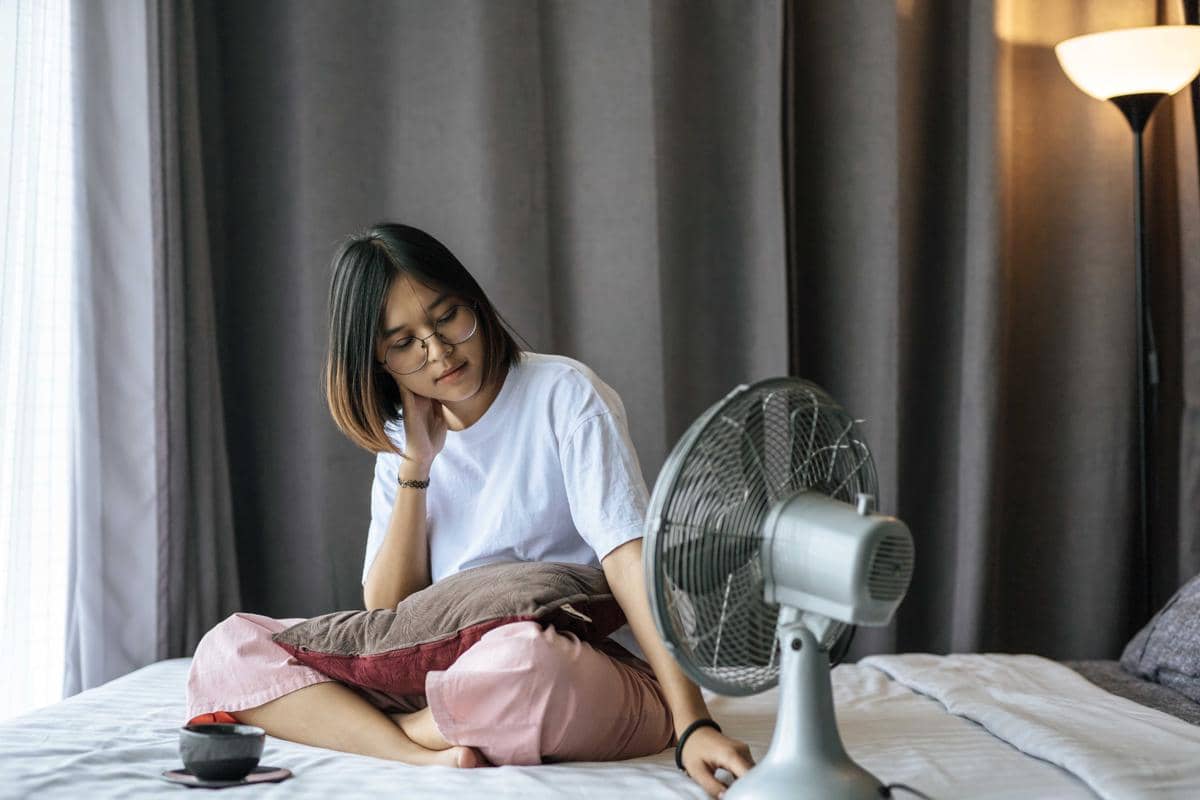 Seorang perempuan duduk di atas tempat tidur sambil menatap kipas angin yang menyala di depannya di dalam kamar dengan tirai abu-abu.