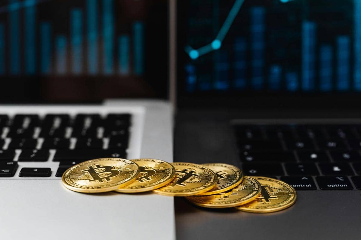 Beberapa koin emas bitcoin diletakkan di antara dua laptop dengan layar menampilkan grafik keuangan berwarna biru.