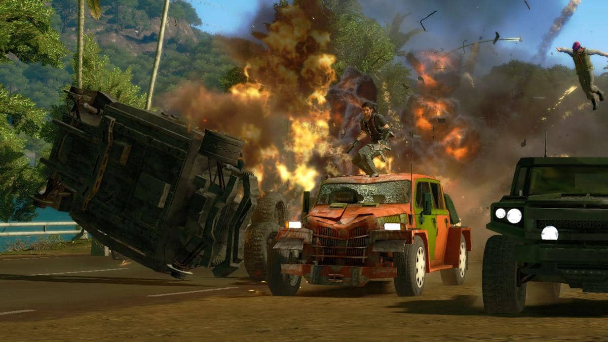 Adegan aksi dalam permainan Just Cause 2 menampilkan karakter berdiri di atas mobil merah saat ledakan besar terjadi di jalan.