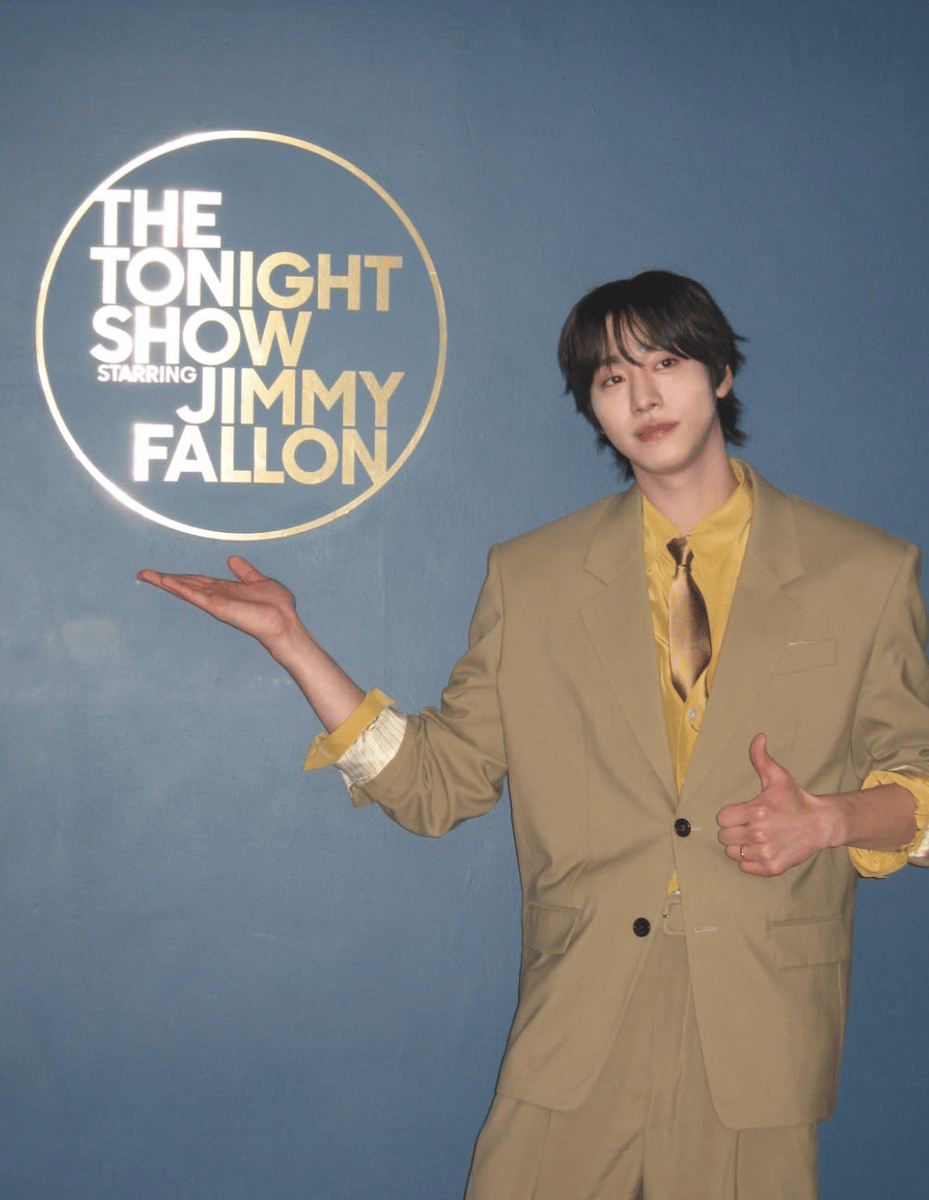 Seorang pria mengenakan setelan jas berwarna krem dan kemeja kuning berpose di depan logo The Tonight Show Starring Jimmy Fallon.