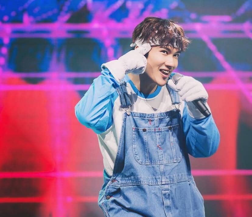 Seorang pria mengenakan baju biru dan overall denim sedang bernyanyi di atas panggung dengan latar cahaya warna-warni.