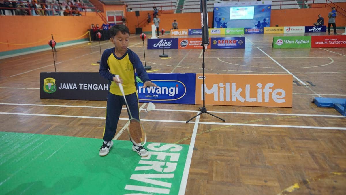 Seorang anak mengenakan seragam olahraga biru kuning sedang berlatih bulu tangkis di lapangan indoor dengan berbagai spanduk sponsor di sekitarnya.