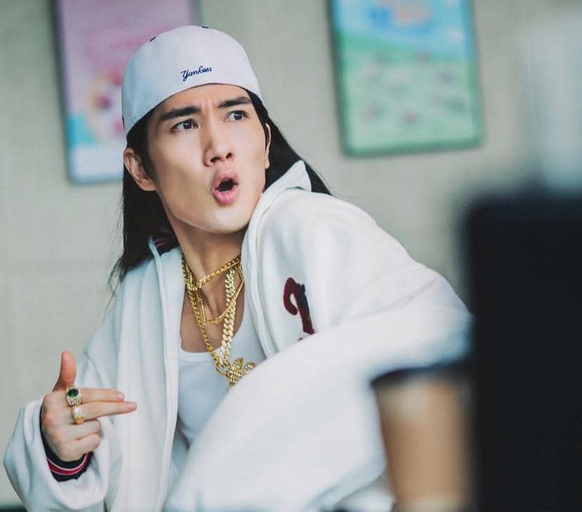 Seorang pria mengenakan jaket putih, topi terbalik, dan rantai emas sedang berpose ekspresif di dalam ruangan.