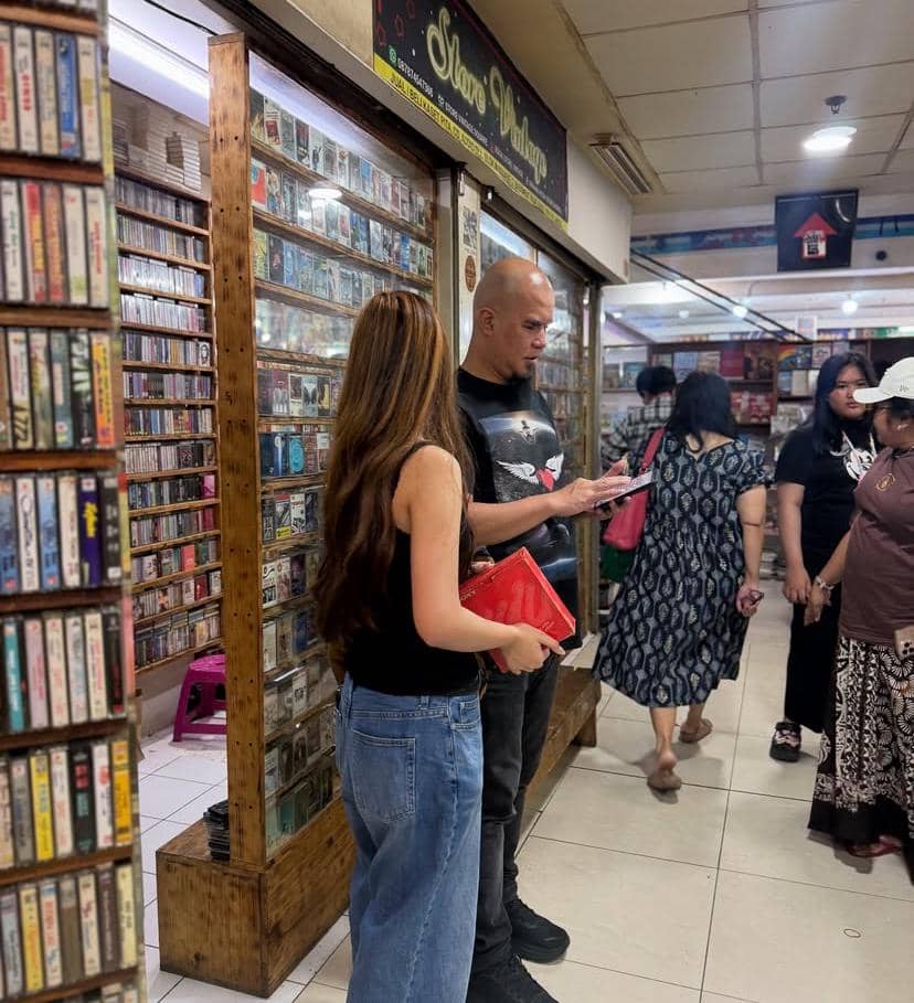 Seorang pria dan seorang gadis berdiri di depan toko kaset musik di dalam pusat perbelanjaan, sambil melihat koleksi kaset yang dipajang.