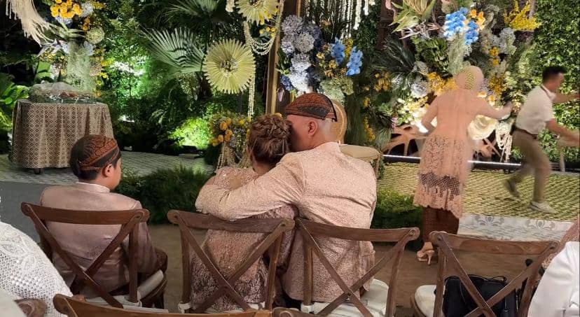 Seorang pria memeluk seorang anak perempuan di acara pesta dengan dekorasi bunga warna-warni dan pencahayaan hangat di malam hari.