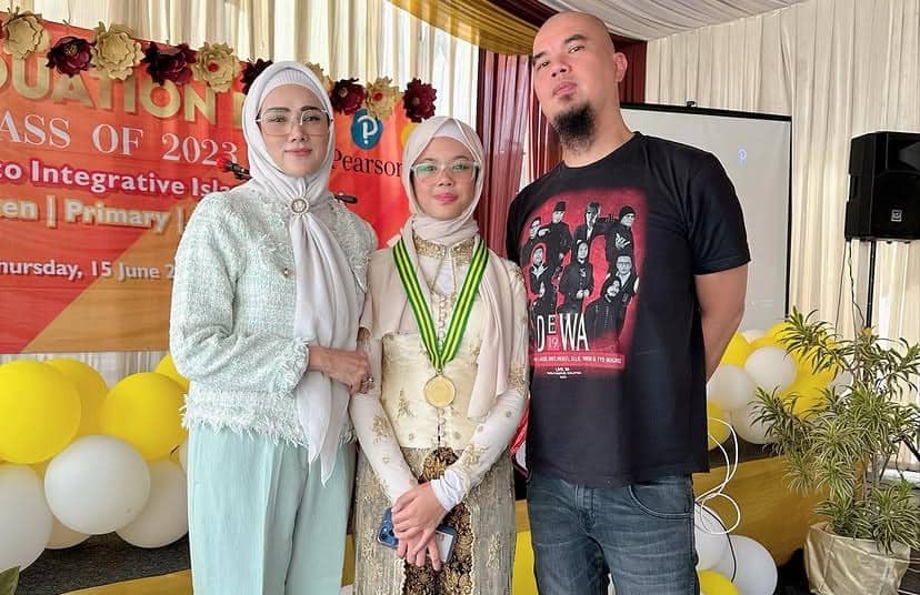 Seorang anak perempuan mengenakan medali kelulusan berpose bersama dua orang dewasa di acara wisuda sekolah dengan dekorasi balon dan spanduk.