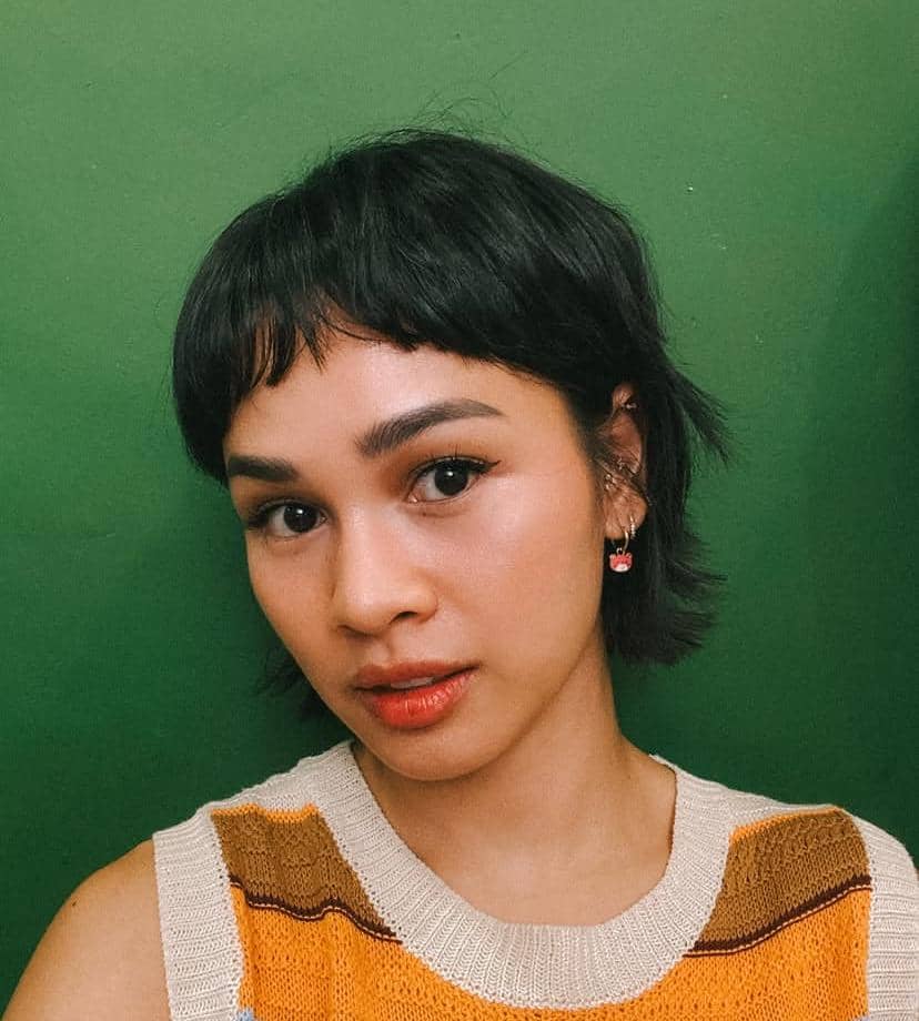 Seorang perempuan berambut pendek hitam mengenakan atasan rajut oranye bermotif garis dengan latar belakang hijau polos.