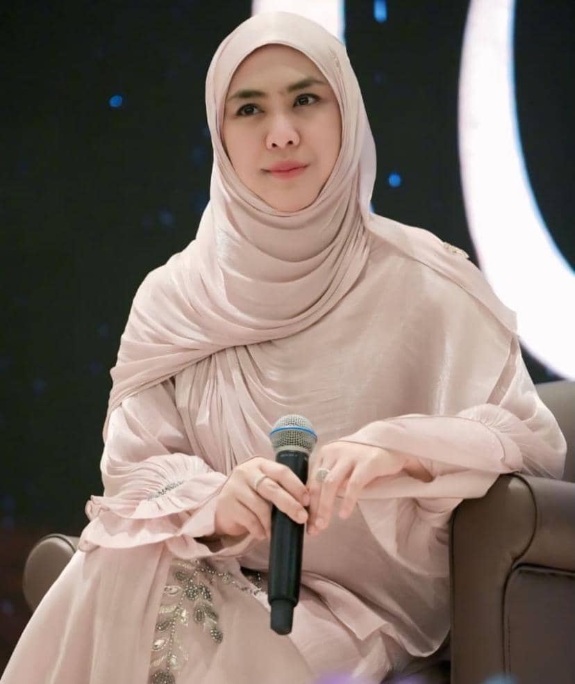 Seorang perempuan berhijab warna pastel duduk di kursi sambil memegang mikrofon dengan latar belakang gelap bertabur cahaya.
