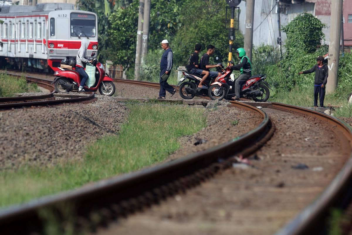 Beberapa pengendara motor dan pejalan kaki melintasi rel kereta api saat kereta mendekat di area perlintasan tanpa palang pintu.