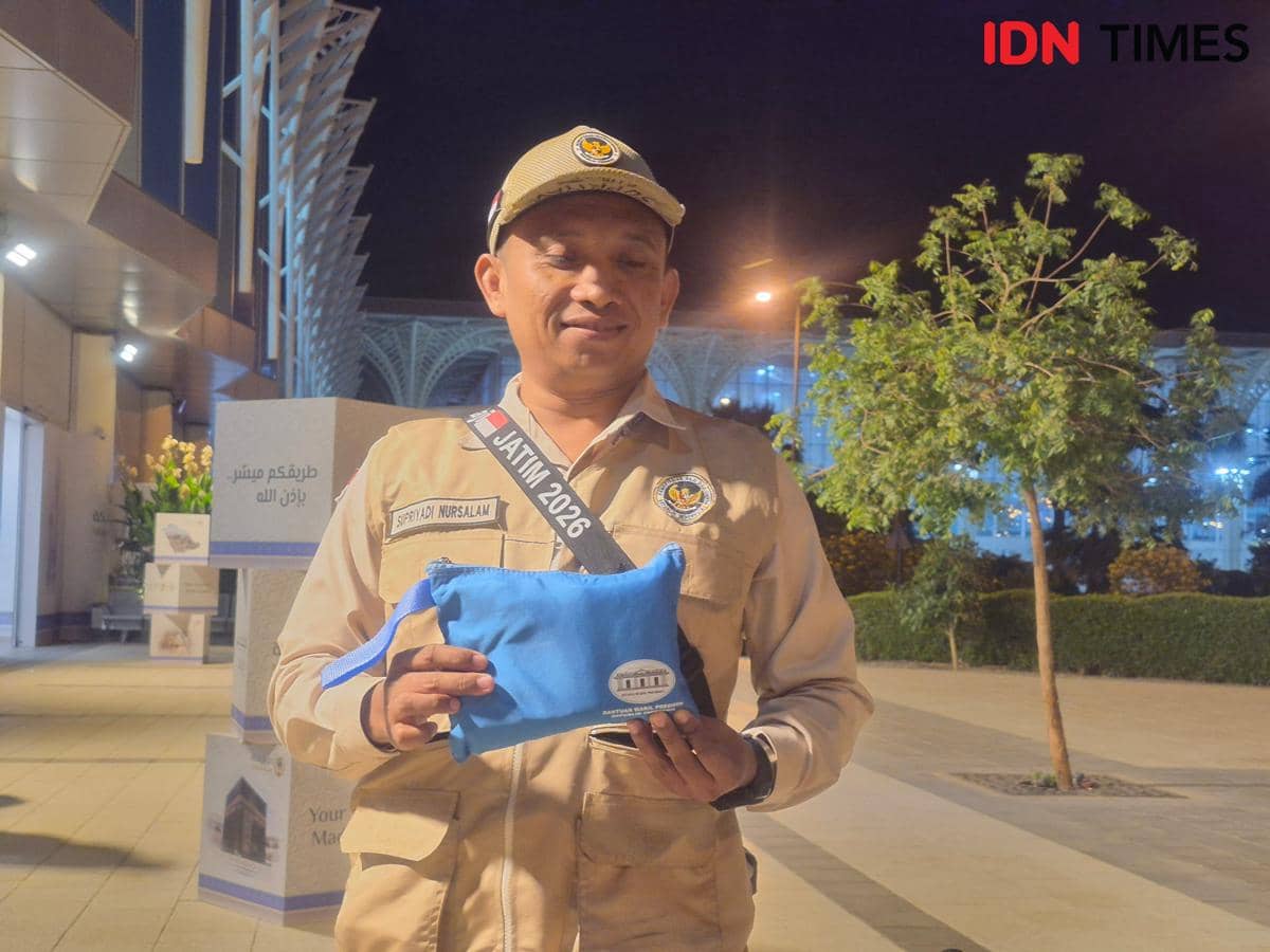 Seorang pria berseragam krem memegang travel kit berwarna biru bertuliskan logo resmi di area luar gedung pada malam hari.