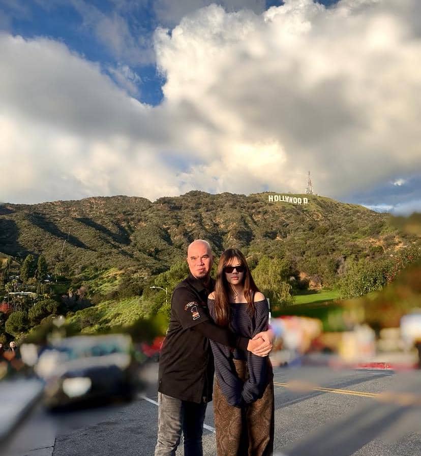 Seorang pria dan seorang gadis berpose bersama di jalan dengan latar belakang bukit bertuliskan Hollywood di bawah langit cerah berawan.
