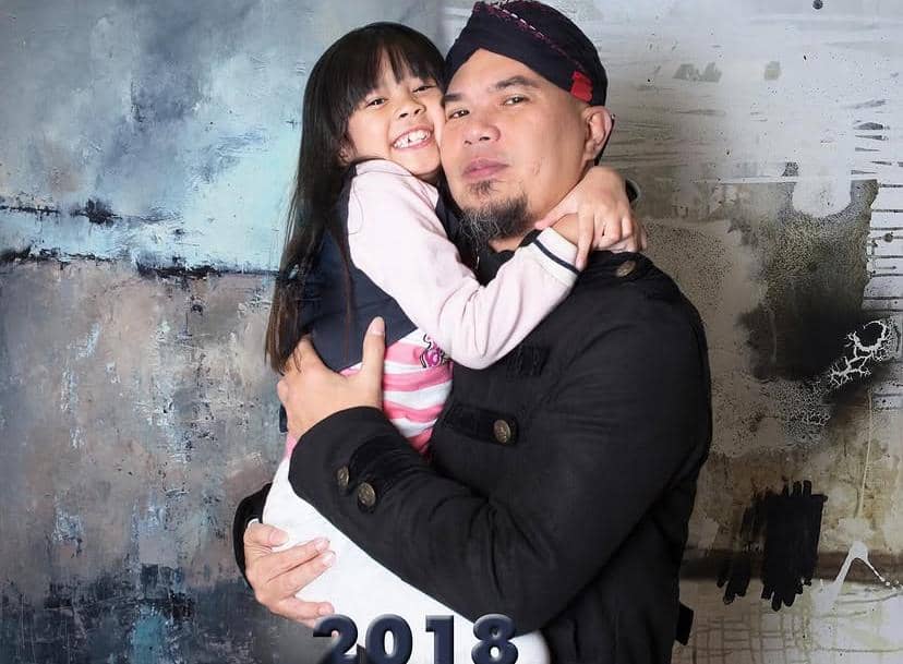 Seorang pria mengenakan pakaian hitam dan ikat kepala merah hitam memeluk seorang anak perempuan yang tersenyum lebar di depannya.