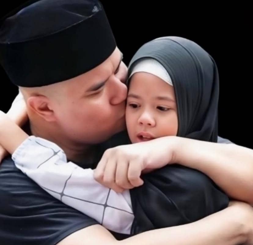 Seorang pria mengenakan peci hitam memeluk dan mencium pipi seorang anak perempuan berhijab abu-abu dengan latar belakang hitam.