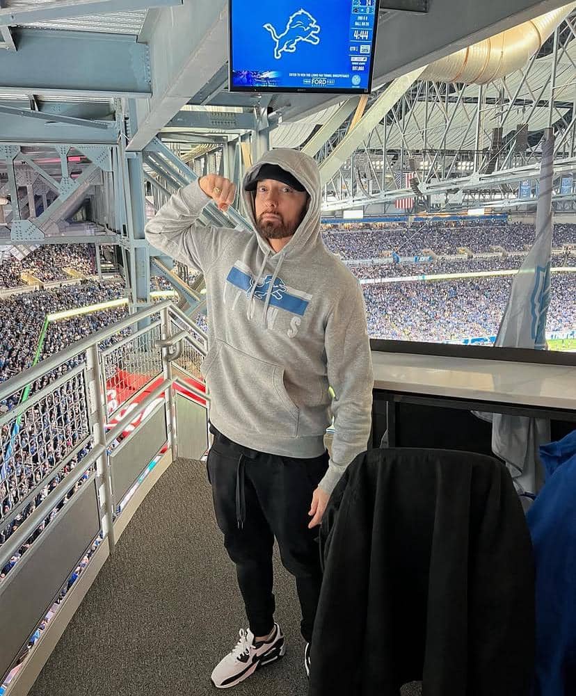 Seorang pria mengenakan hoodie abu-abu dan topi hitam berdiri di tribun stadion dengan latar belakang pertandingan sepak bola Amerika.