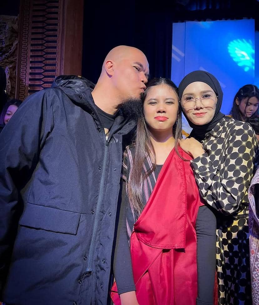Seorang pria berkepala plontos mencium kepala seorang gadis muda di tengah, sementara seorang wanita berhijab berdiri di samping mereka dengan senyum hangat.