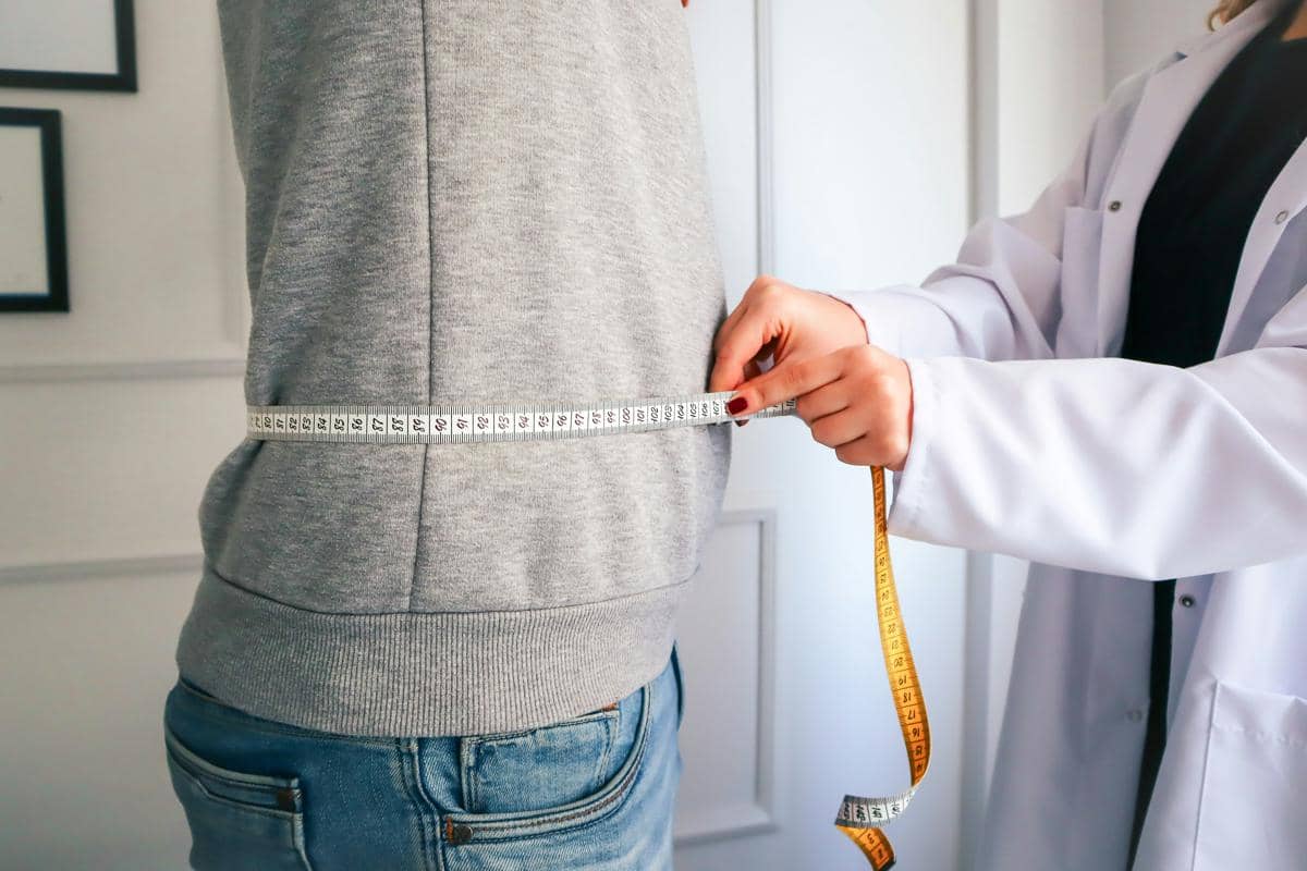 Dokter mengenakan jas putih sedang mengukur lingkar perut pasien dengan pita ukur di ruangan pemeriksaan medis.